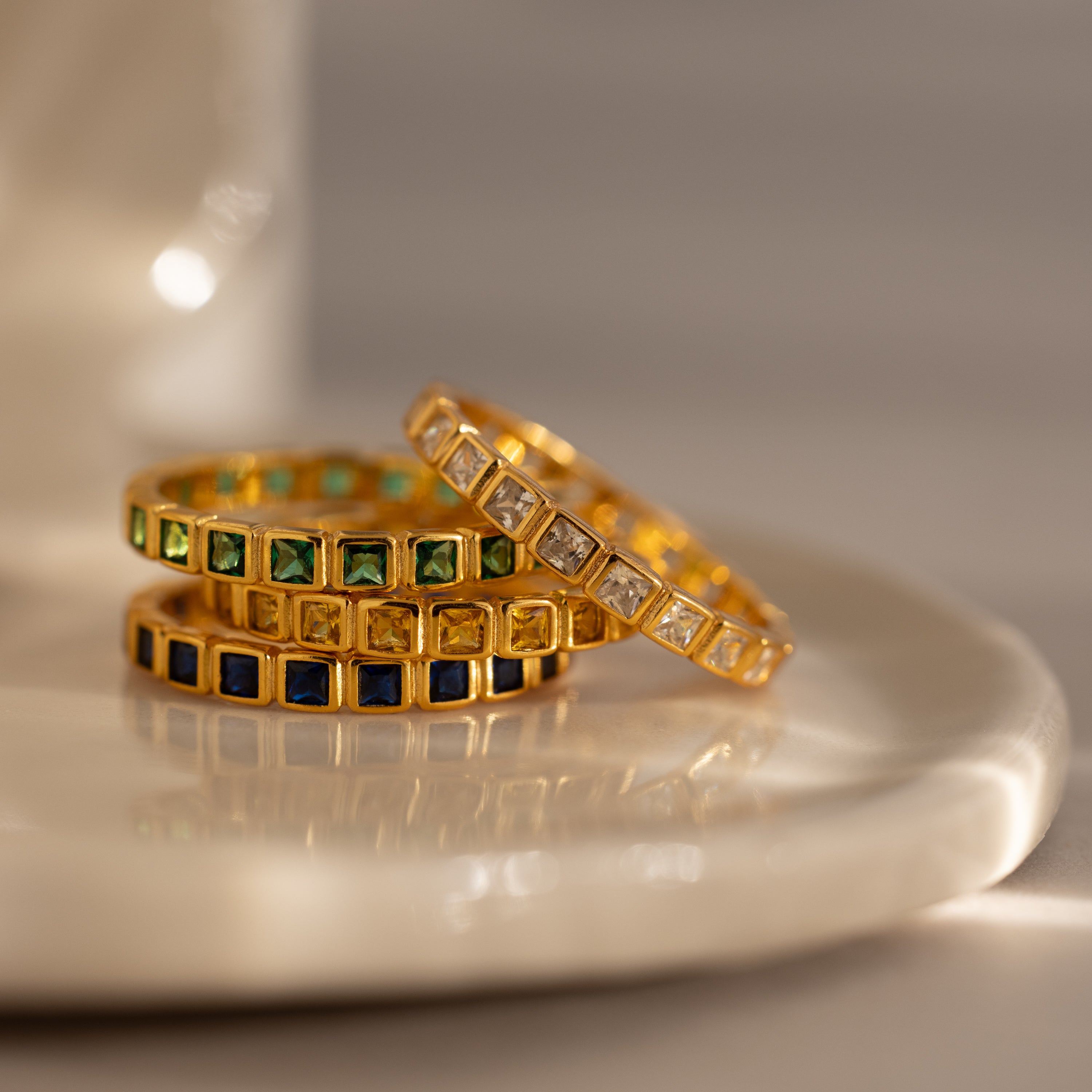 Three Eternity Bezel Sapphire Rings with square-cut green, blue, and clear gemstones are stacked on a glossy white surface.