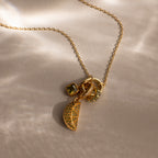 Gold chain necklace with several gold and gemstone charms, including a sparkling pavé Peridot Lime Charm, displayed on a beige surface with soft shadows.