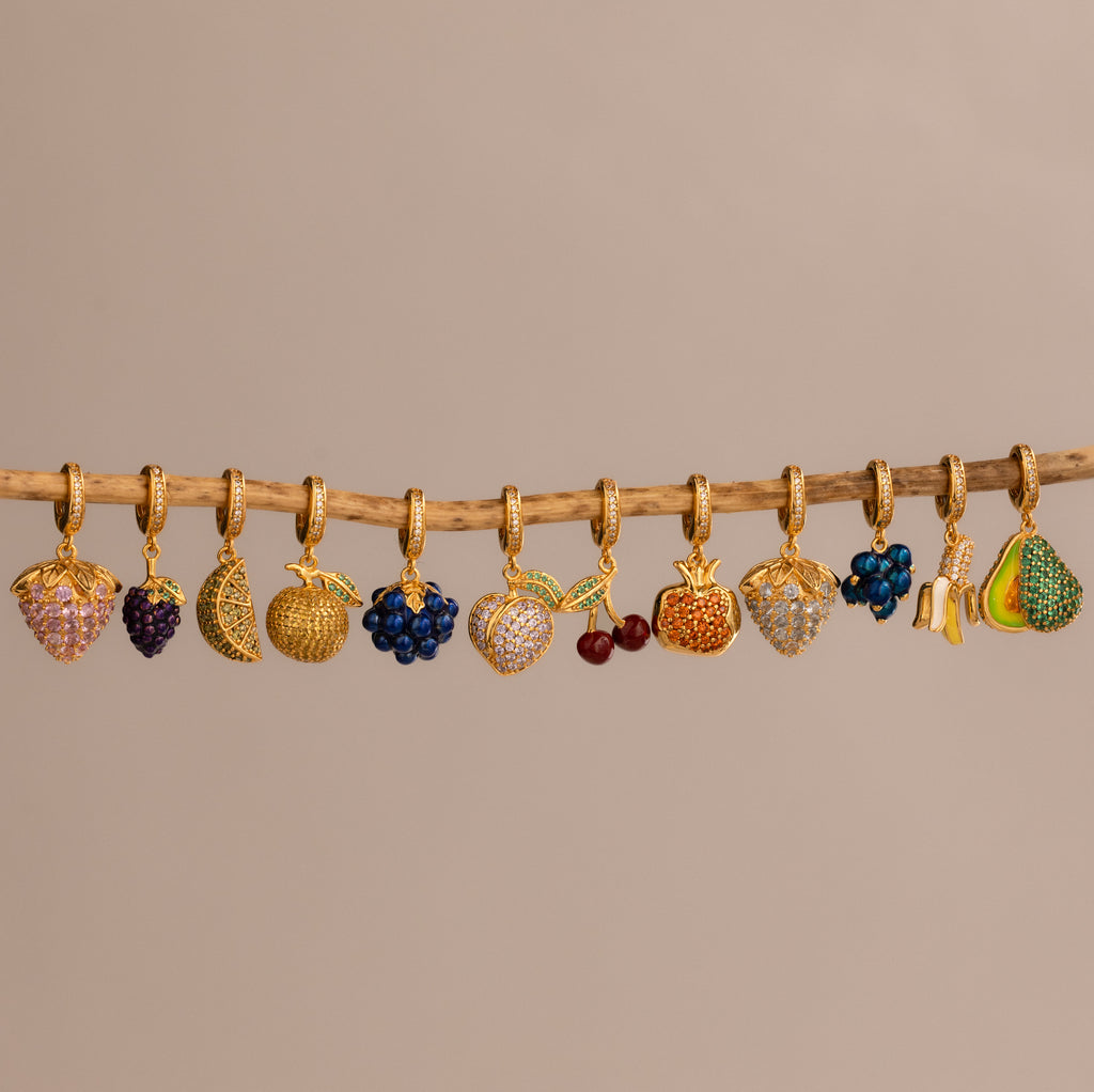 The Emerald Avocado Charm, shaped like a gold fruit and adorned with pavé stones, hangs on a wooden stick against a plain beige background, making it a sparkling addition to any jewelry collection.
