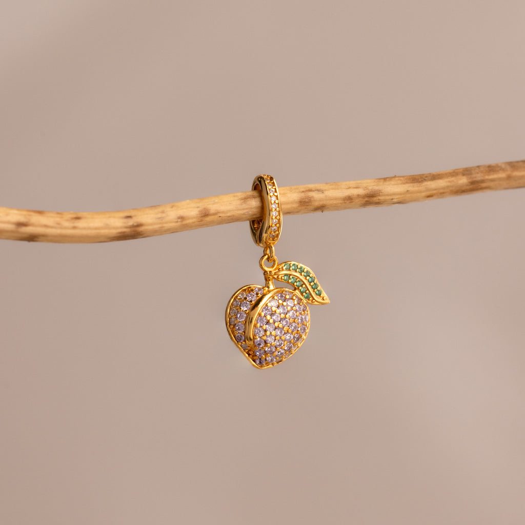 The Alexandrite Peach Charm features a gold peach-shaped pendant with pavé stones, suspended from a branch on a beige background—a delightful addition to any jewelry collection.