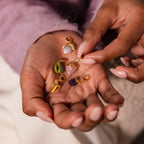 A hand holds six vibrant gold charms from a jewelry collection while another hand picks up the sparkling Emerald Avocado Charm.