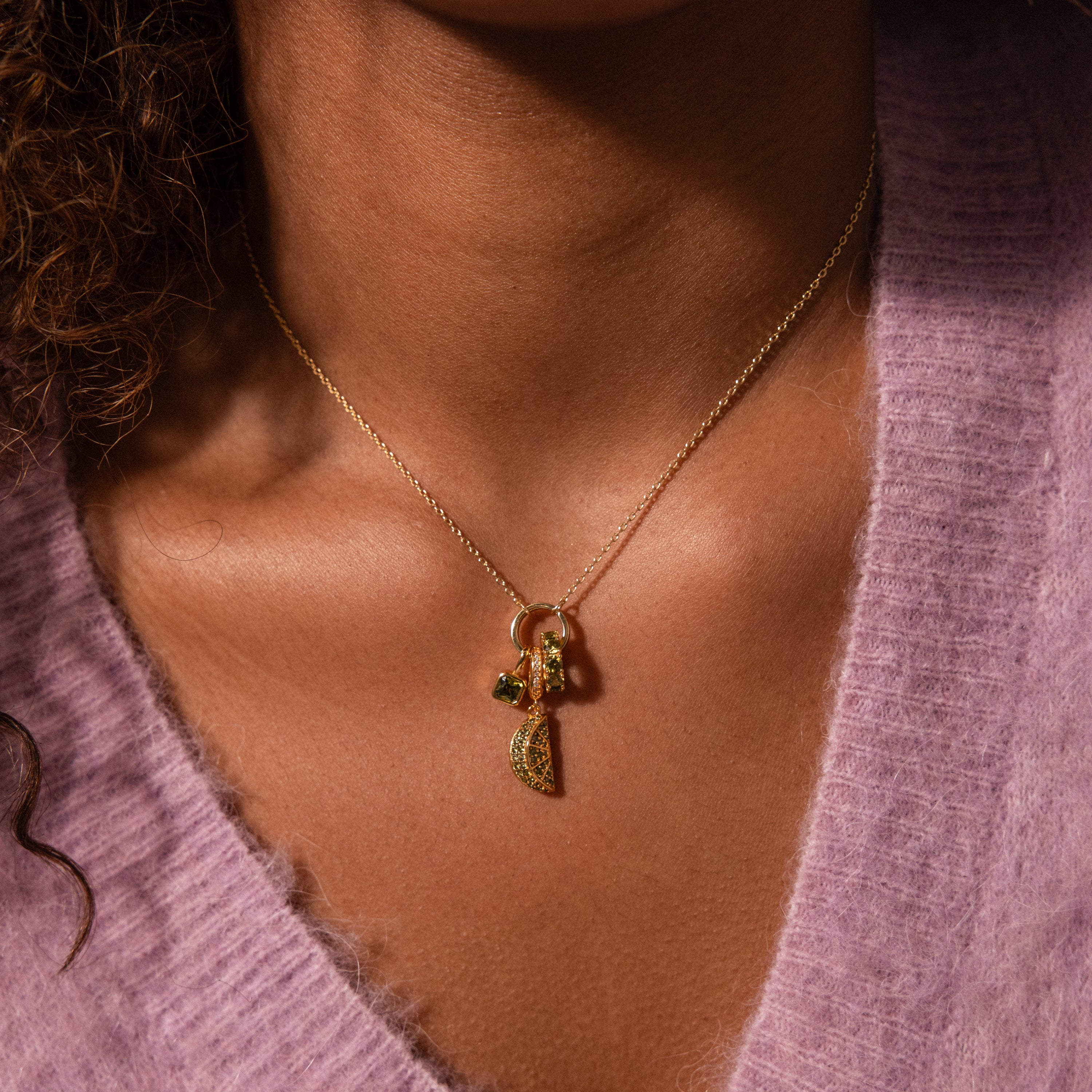 Close-up of the Emerald Avocado Charm gold necklace with pavé stones and fruit-shaped pendants, worn over a soft lavender sweater.