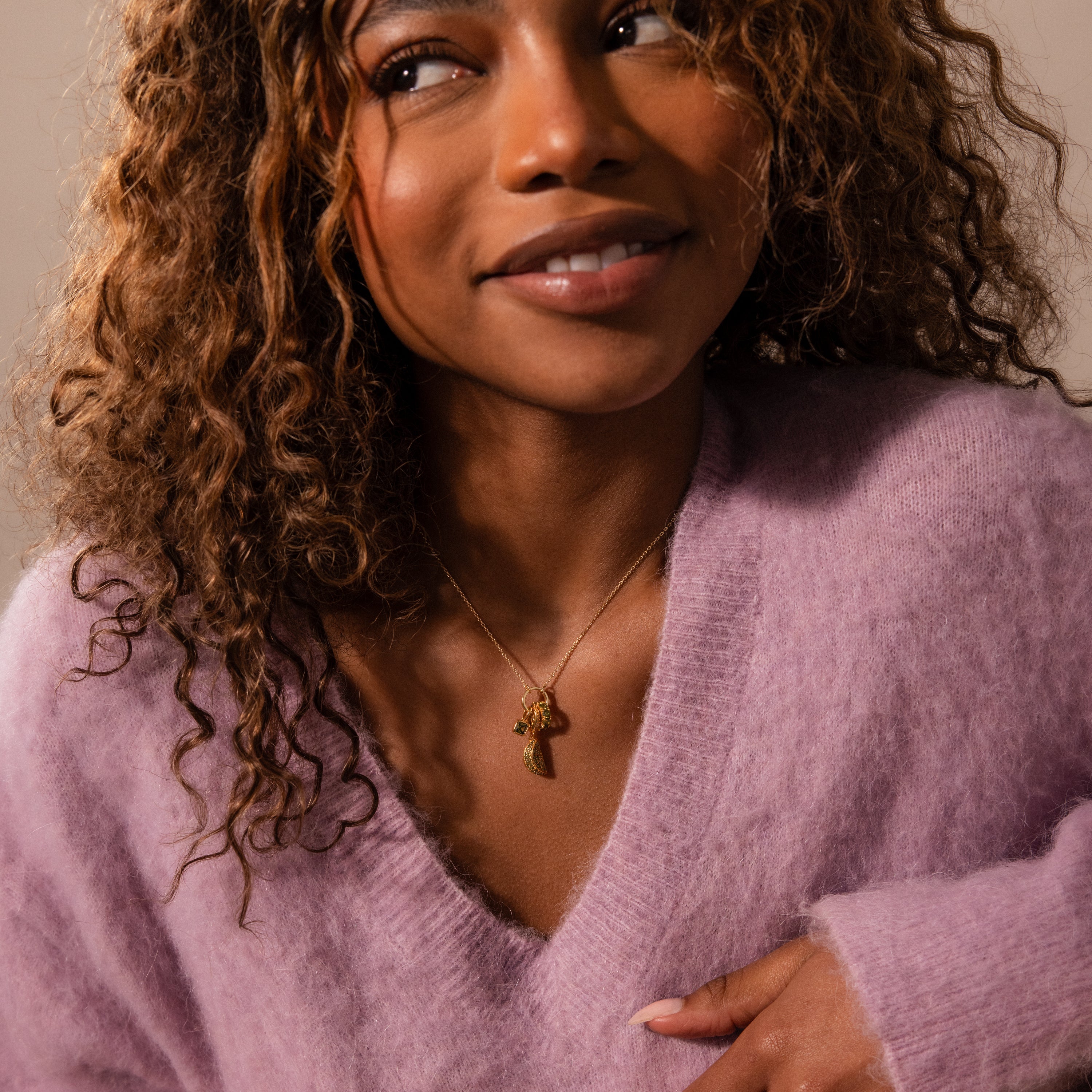 Smiling woman in a fuzzy lavender sweater, wearing a gold necklace featuring the Sapphire Blackberry Charm from the jewelry collection, adorned with pavé stones.