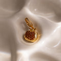 The Garnet Pomegranate Charm features red pavé stones set in a gold pendant, elegantly displayed on cream-colored fabric—a charming addition to any jewelry collection.