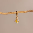 The Diamond Banana Charm, featuring pavé stones in a gold banana shape, hangs elegantly from a thin branch on a beige background—an eye-catching addition to any jewelry collection.