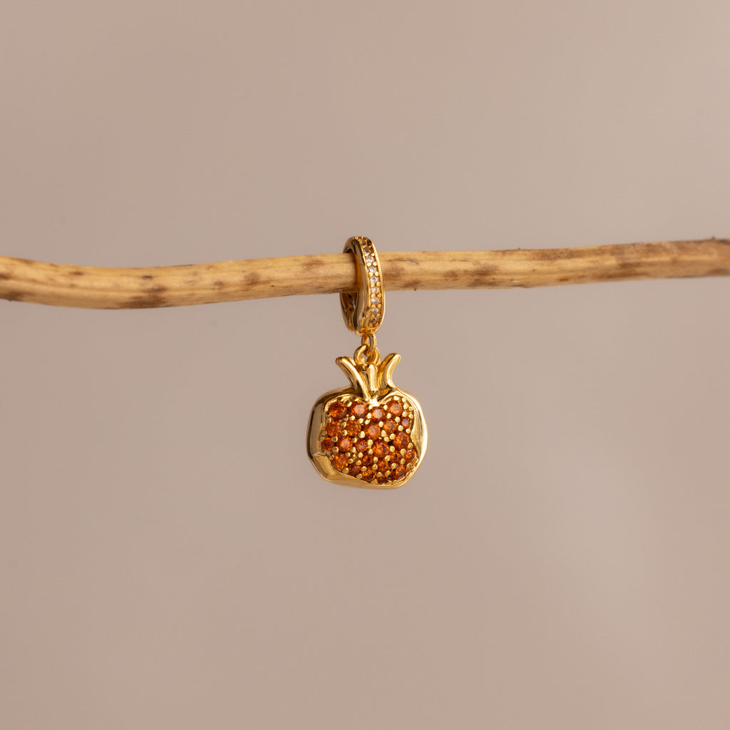 The Garnet Pomegranate Charm, featuring red pavé stones, hangs from a branch against a neutral background.