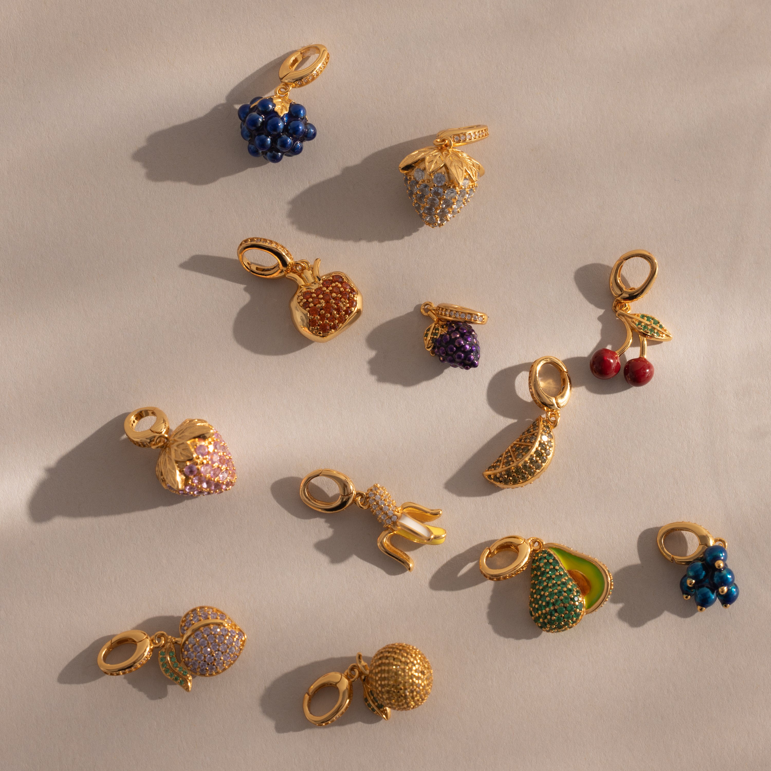 The Emerald Avocado Charm, adorned with vivid stones and shaped like a fruit, rests elegantly on a beige surface with soft shadows—a refined addition to any jewelry collection.