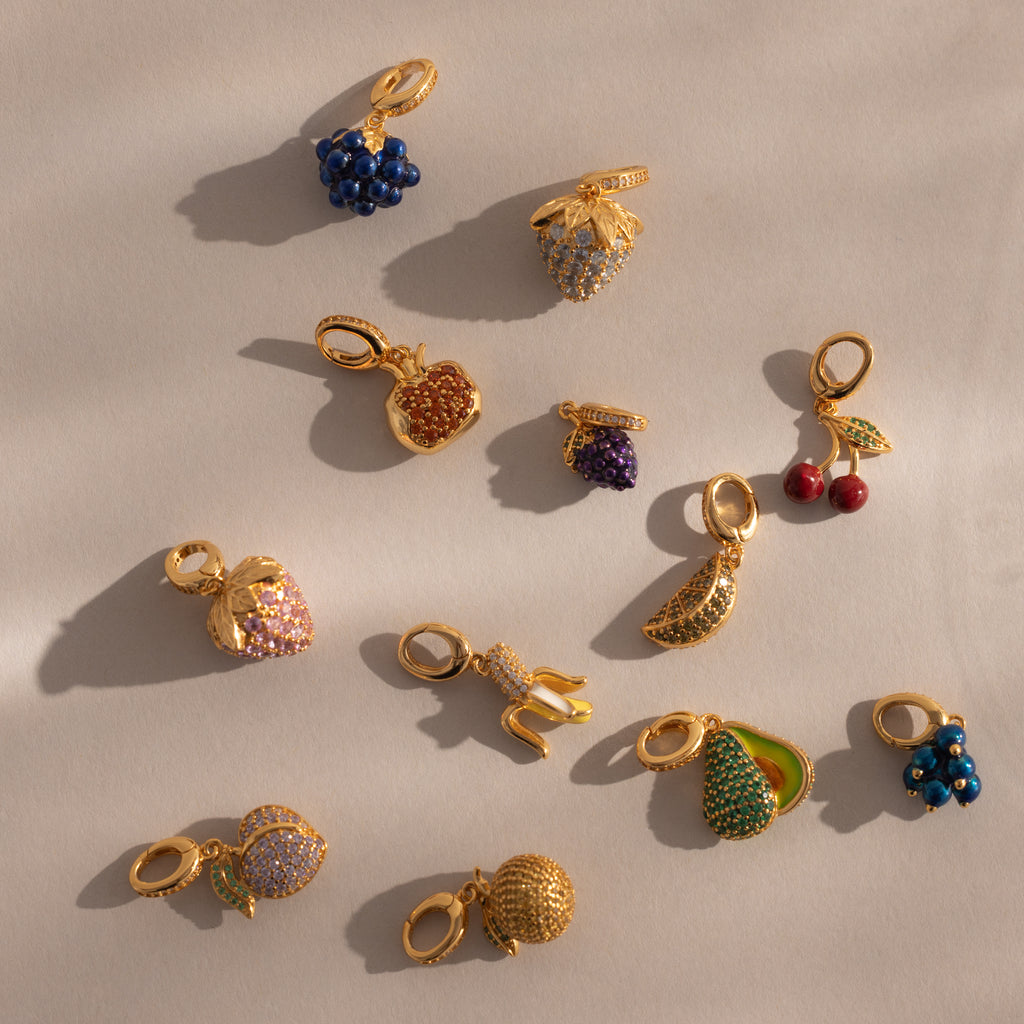The Sapphire Blackberry Charm from the Birthstone Fruit Charm jewelry collection, accented with pavé stones and shaped like a blackberry, is displayed on a beige surface.