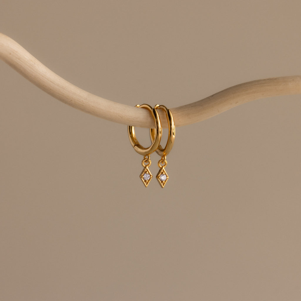The Ericka Birthstone Huggies in Diamond / April hang from a curved beige branch on a neutral background—an elegant choice for anyone who loves diamond-accented or personalized earrings.