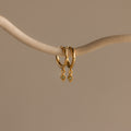 Two Ericka Birthstone Huggies in Peridot (August) hang from a curved, light-colored branch—elegant hoops with sparkling charms that add a vibrant touch to any style.