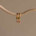 The Ericka Ruby Huggies are gold hoop earrings featuring small red diamond-shaped pendants, elegantly displayed on a curved beige branch against a neutral background.