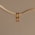 The Ericka Ruby Huggies are gold hoop earrings featuring small red diamond-shaped pendants, elegantly displayed on a curved beige branch against a neutral background.