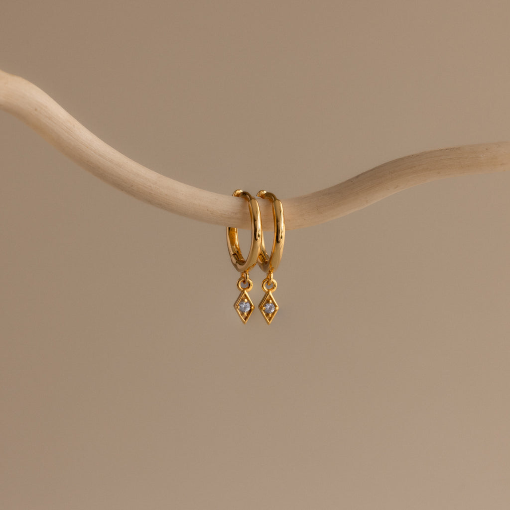 Ericka Birthstone Huggies in Aquamarine (March) hang on a curved beige branch against a neutral background, highlighting elegant aquamarine earrings perfect for any occasion.