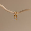 Ericka Birthstone Huggies in Aquamarine (March) hang on a curved beige branch against a neutral background, highlighting elegant aquamarine earrings perfect for any occasion.