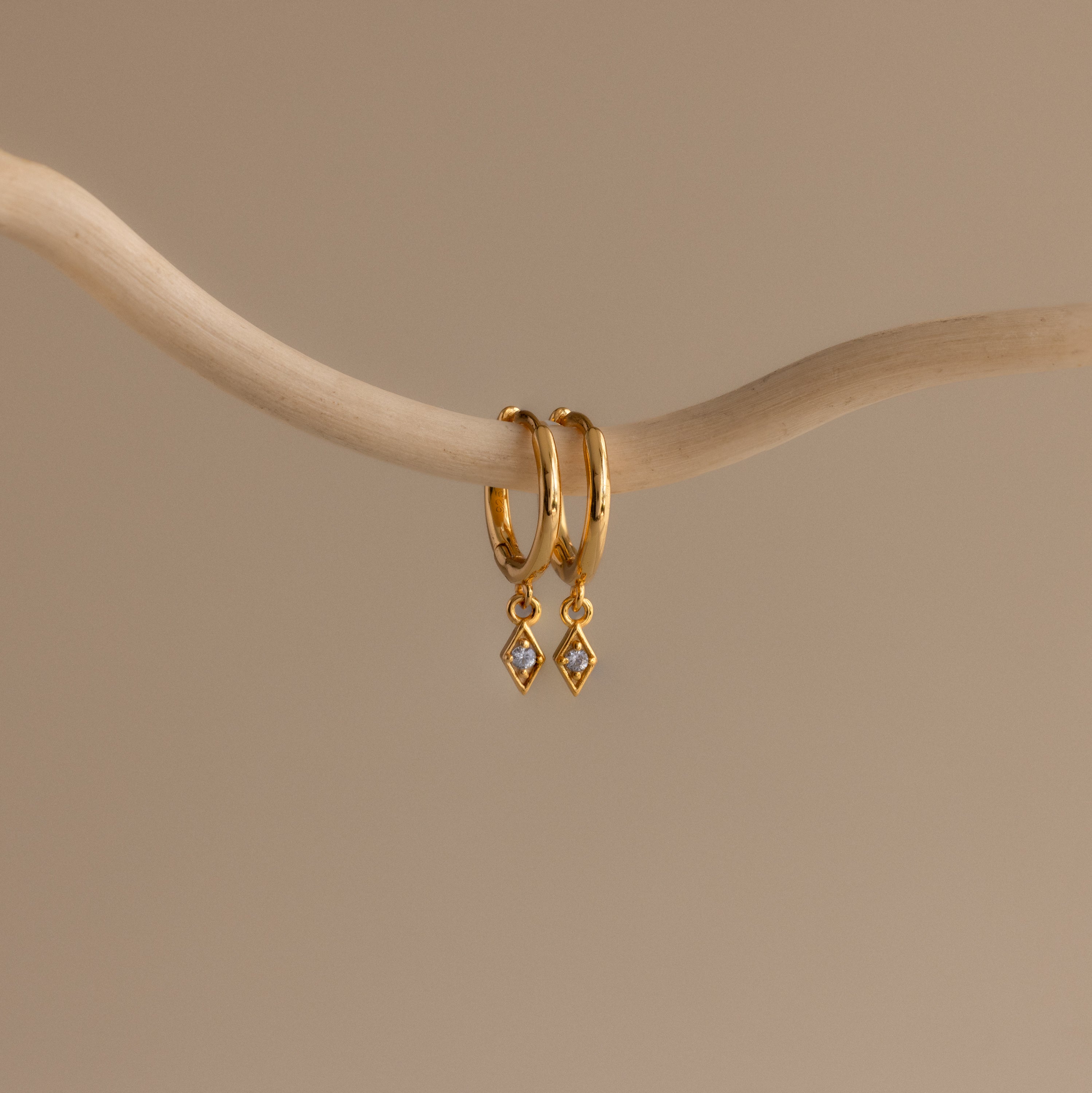 Ericka Birthstone Huggies in Aquamarine (March) hang on a curved beige branch against a neutral background, highlighting elegant aquamarine earrings perfect for any occasion.