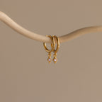 Ericka Alexandrite Huggies evoke the allure of diamond charm earrings, elegantly hanging from a smooth, curved beige branch against a plain background.