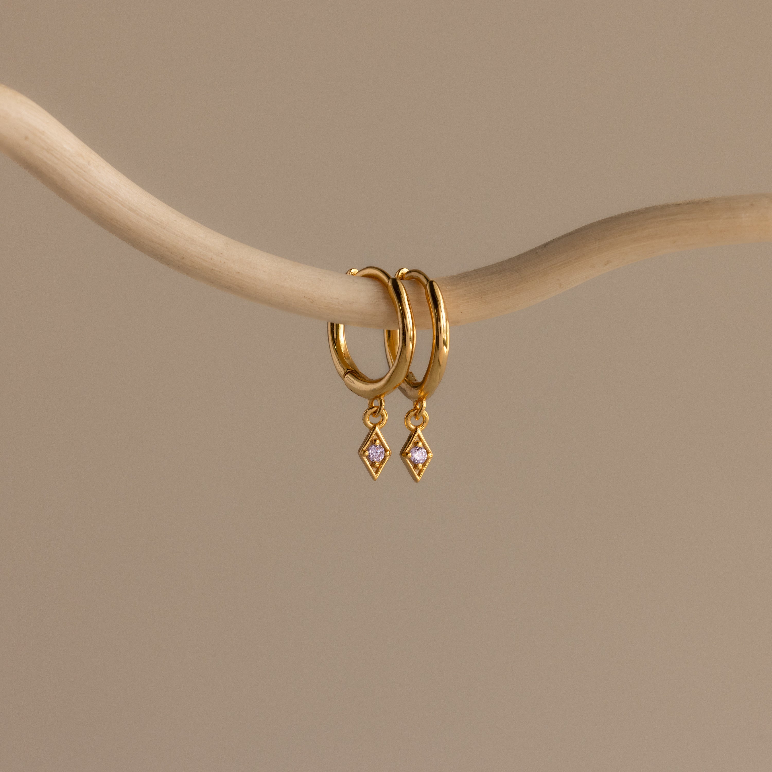 Ericka Alexandrite Huggies evoke the allure of diamond charm earrings, elegantly hanging from a smooth, curved beige branch against a plain background.