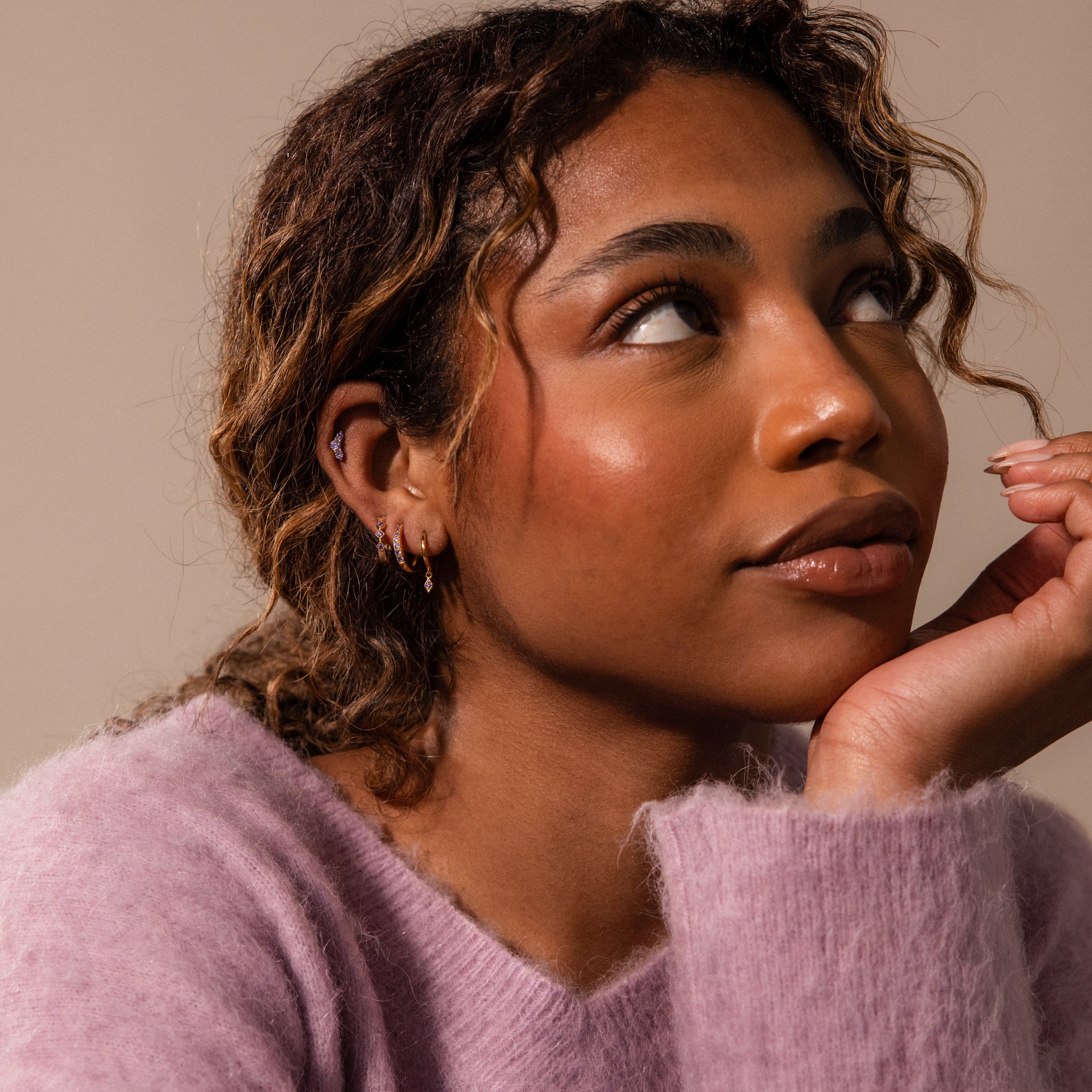 A woman with wavy hair and hoop earrings, wearing a fuzzy lavender sweater, gazes thoughtfully upward—her Ericka Aquamarine Huggies add a personalized touch to her elegant look.