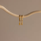 Two Ericka Topaz Huggies earrings, each adorned with a small diamond-shaped charm, hang gracefully from a curved, light-colored branch.