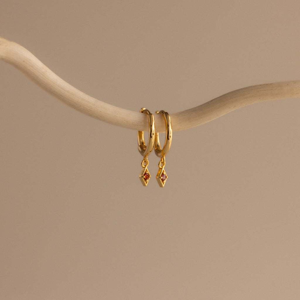 The Ericka Birthstone Huggies in Garnet (January) feature two gold hoops with small diamond-shaped red charms, hanging from a curved beige branch—ideal as birthstone huggies or a unique twist on diamond charm earrings.