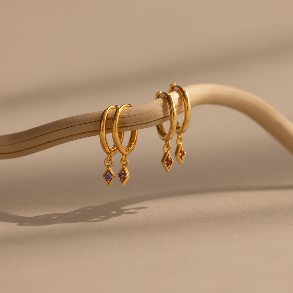 Ericka Amethyst Huggies—gold hoop earrings with amethyst charm pendants—hang on a curved twig against a beige background, offering the perfect touch for personalized jewelry lovers seeking elegant birthstone huggies.