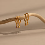 Ericka Amethyst Huggies—gold hoop earrings with amethyst charm pendants—hang on a curved twig against a beige background, offering the perfect touch for personalized jewelry lovers seeking elegant birthstone huggies.