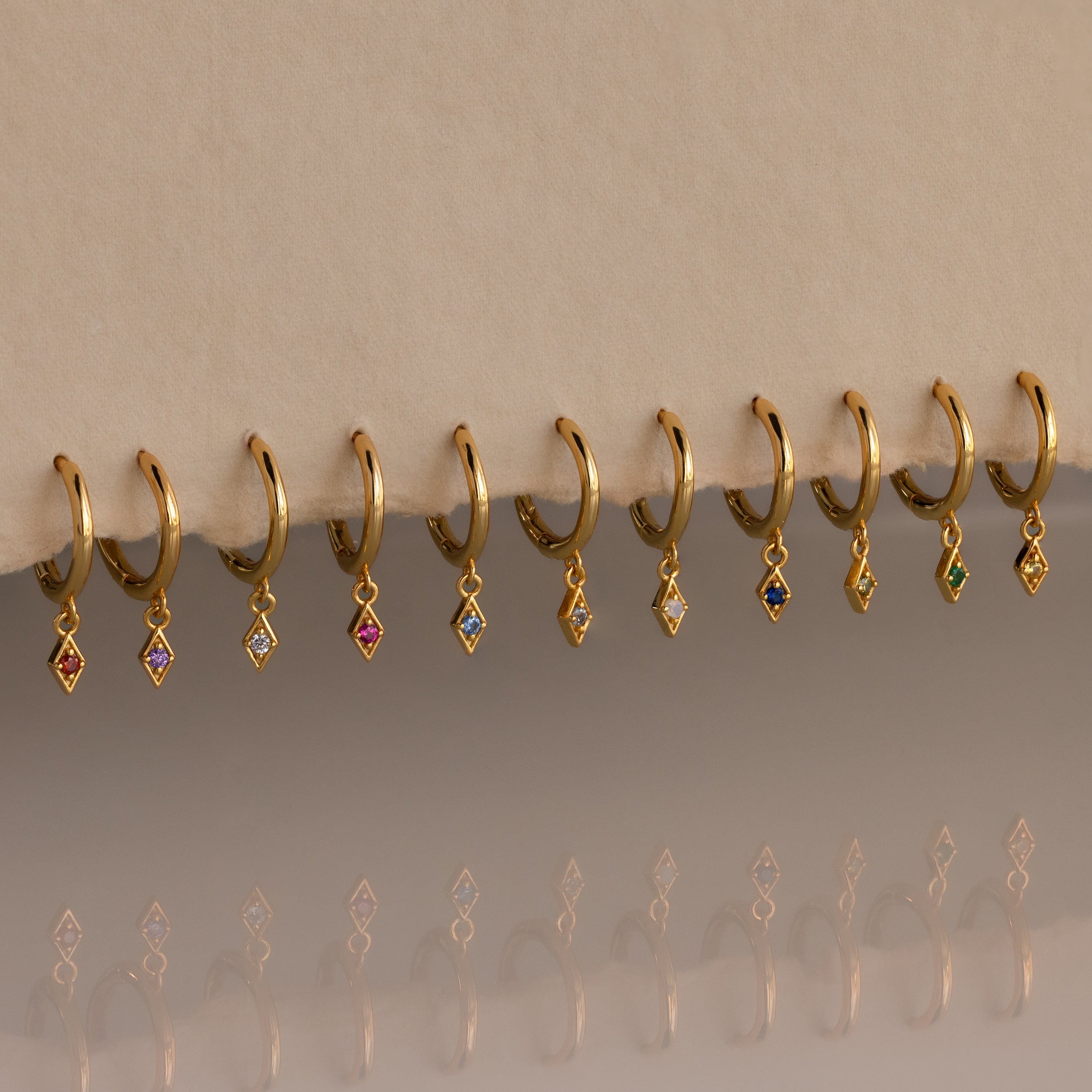 Ten Ericka Birthstone Huggies in Alexandrite (June) rest on a beige surface, their diamond-shaped charms sparkling with colorful reflections—perfect for those who love personalized jewelry.