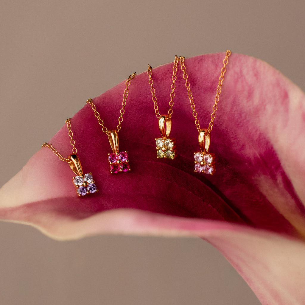 Four gold necklaces with colorful gemstone pendants, including the Ombre Alexandrite Flower Necklace, are displayed elegantly on a pink and white flower petal.