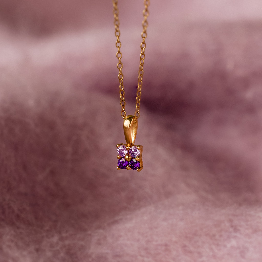 The Ombre Amethyst Flower Necklace features a gold chain with a small flower pendant adorned with purple amethyst gemstones, photographed on a soft pink background.