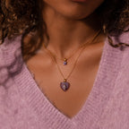 Woman wearing a soft purple sweater and two gold necklaces—one with a heart charm, the other is the Ombre Topaz Flower Necklace featuring a small square pendant.