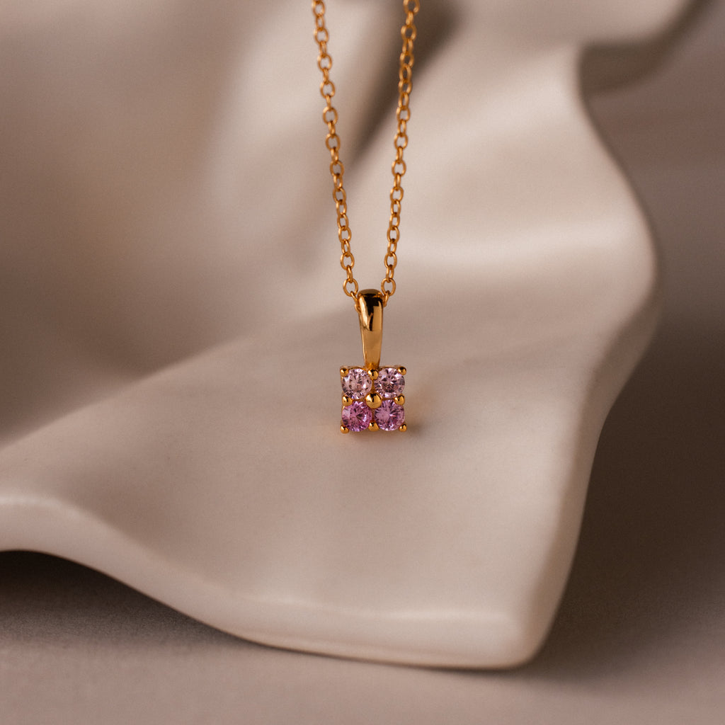 The Ombre Pink Tourmaline Flower Necklace features a gold square pendant adorned with pink and clear gemstones, capturing delicate charm, and is displayed on a soft beige surface.