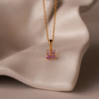 The Ombre Pink Tourmaline Flower Necklace features a gold square pendant adorned with pink and clear gemstones, capturing delicate charm, and is displayed on a soft beige surface.
