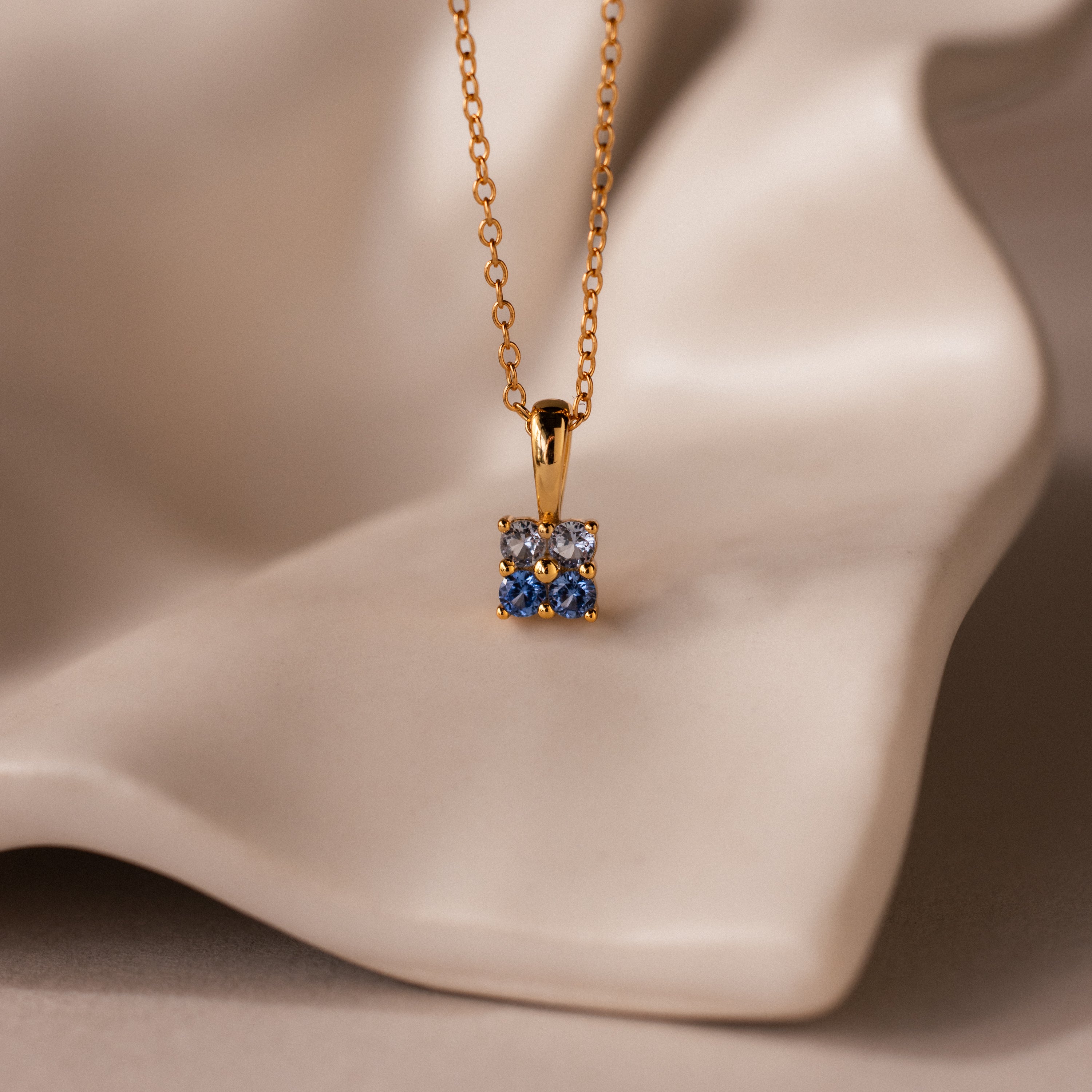 Ombre Aquamarine Flower Necklace featuring a square blue and white pendant, elegantly displayed on a smooth, cream-colored surface.