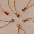 Six gold chain necklaces with colorful gemstone pendants, including the Ombre Emerald Flower Necklace, are arranged in a circular pattern on a beige surface.