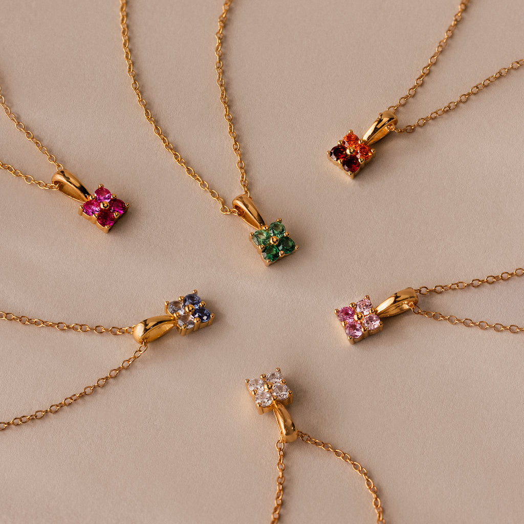 Six Ombre Sapphire Flower Necklaces are arranged in a circle on a beige surface, showcasing their vibrant gradient sapphire pendants.