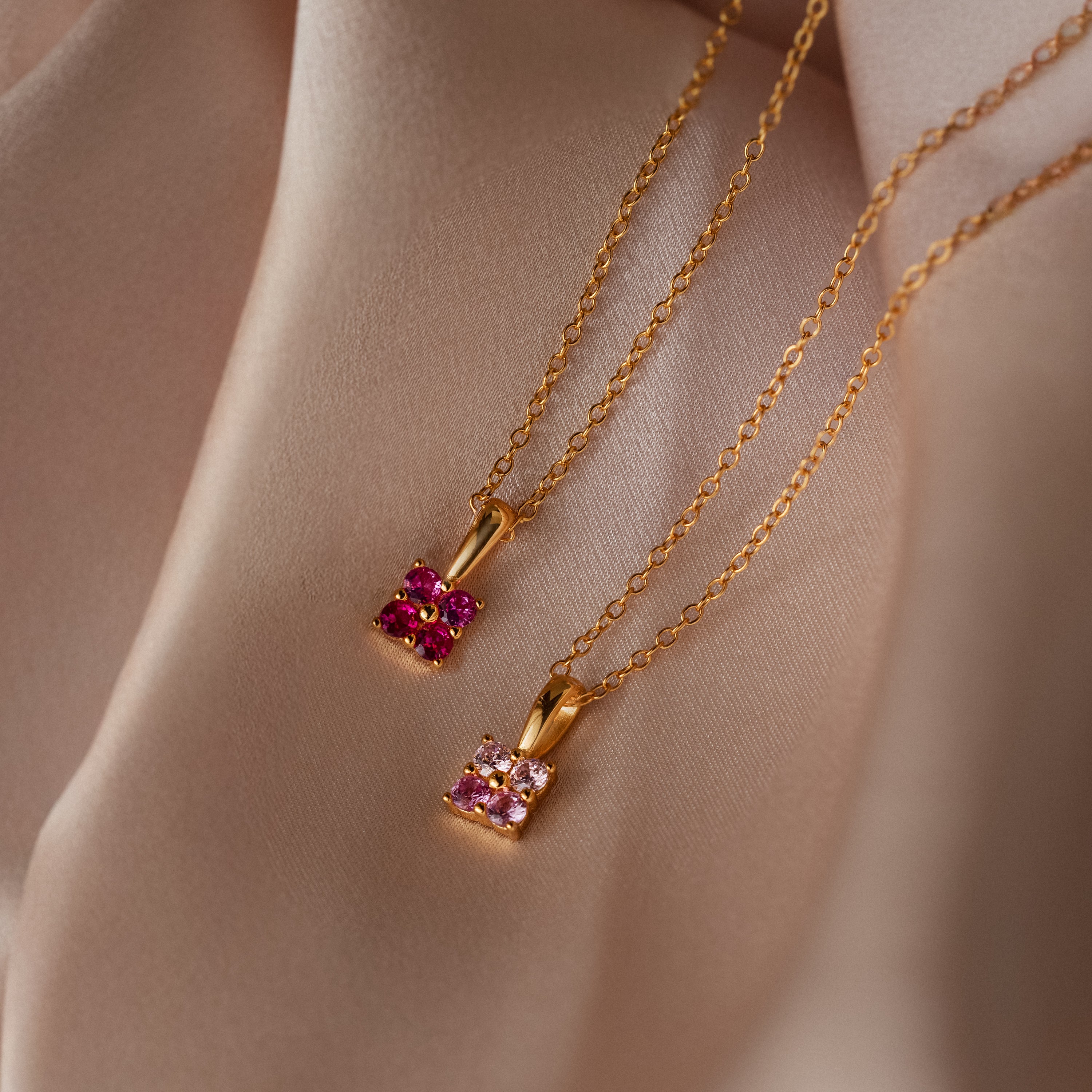 Two gold chain necklaces with pink gemstone pendants rest on soft beige fabric—one is the Ombre Garnet Flower Necklace featuring a delicate ombre birthstone design.