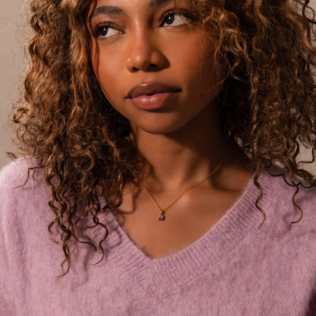 A woman with curly hair wears a light purple sweater and an Ombre Birthstone Flower Necklace featuring a small purple pendant.