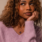 A woman with curly hair in a soft lavender sweater rests her chin on her hand, wearing layered necklaces including the delicate Ruby Crystal Necklace.