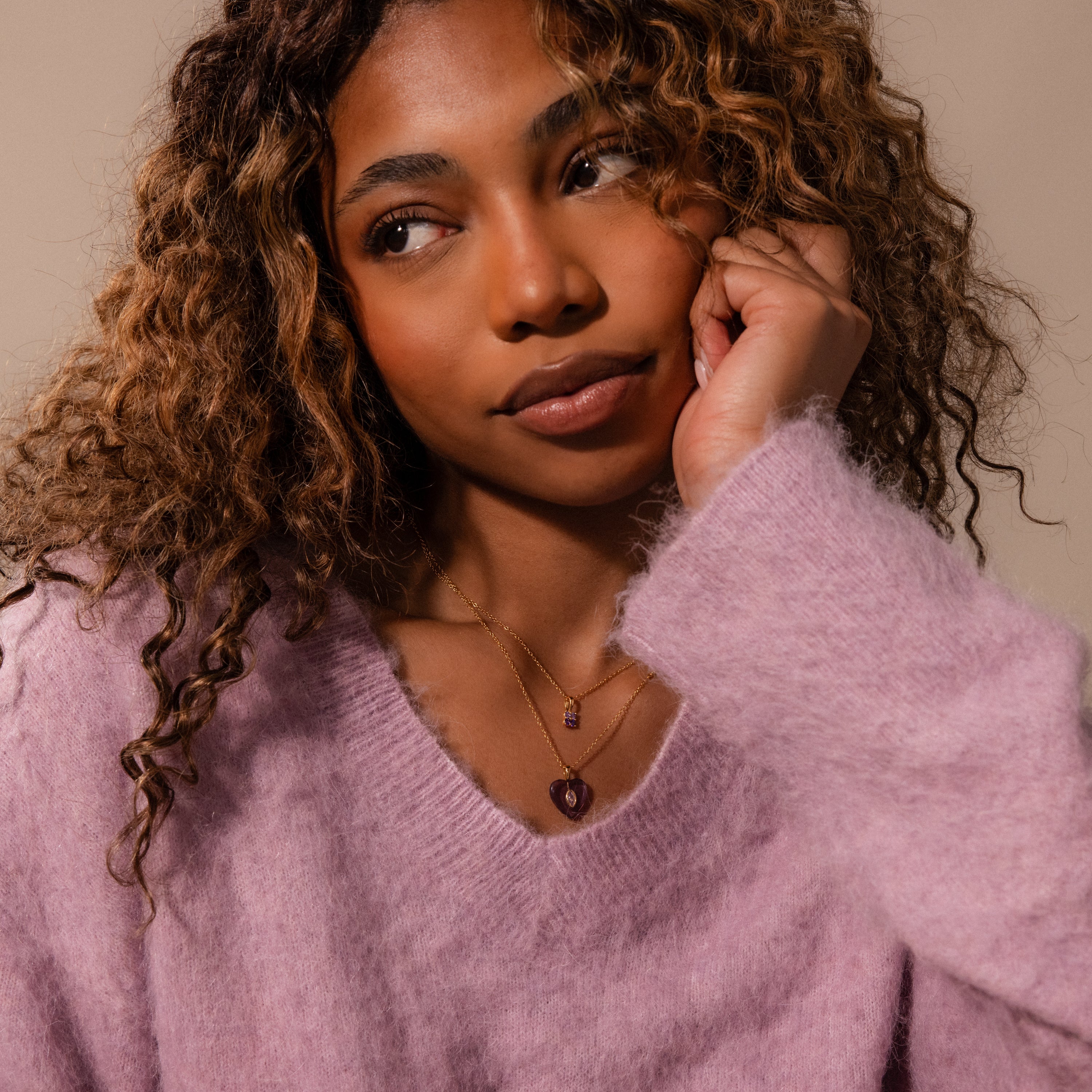 A woman in a fuzzy lavender sweater looks thoughtful, resting her face on her hand, showcasing layered necklaces including a sparkling Amethyst Crystal Necklace.