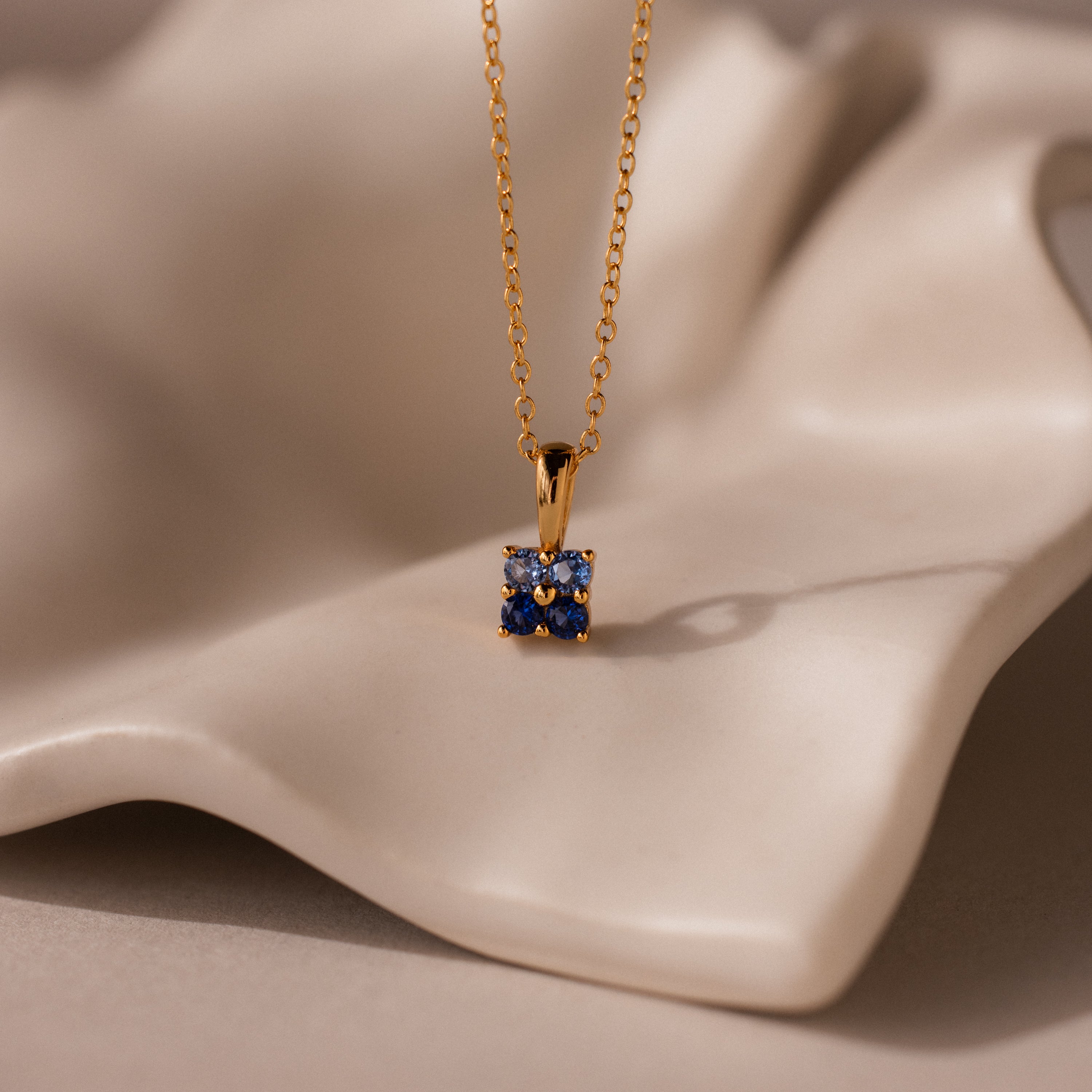 Ombre Blue Zircon Flower Necklace featuring a square blue gemstone pendant displayed on soft, wavy beige fabric—ideal for those who love elegant birthstone jewelry.