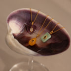 A Peridot Crystal Necklace with a bezel set birthstone and colorful gemstones is displayed on a large purple and white seashell.
