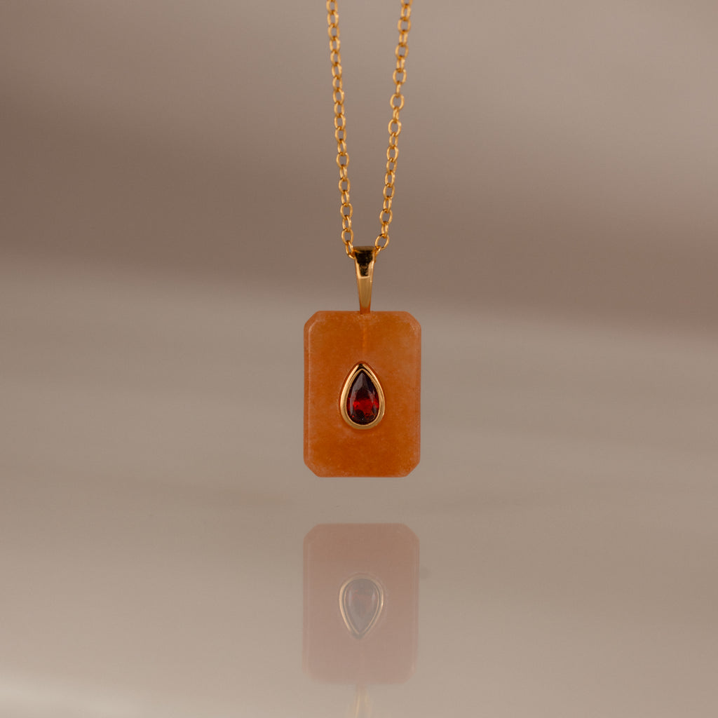 The Garnet Crystal Necklace features a gold chain with a rectangular orange pendant and a teardrop red gemstone, making it a unique birthstone jewelry piece or an eye-catching Birthstone Crystal Necklace.