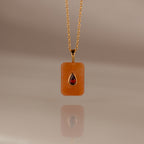 The Garnet Crystal Necklace features a gold chain with a rectangular orange pendant and a teardrop red gemstone, making it a unique birthstone jewelry piece or an eye-catching Birthstone Crystal Necklace.