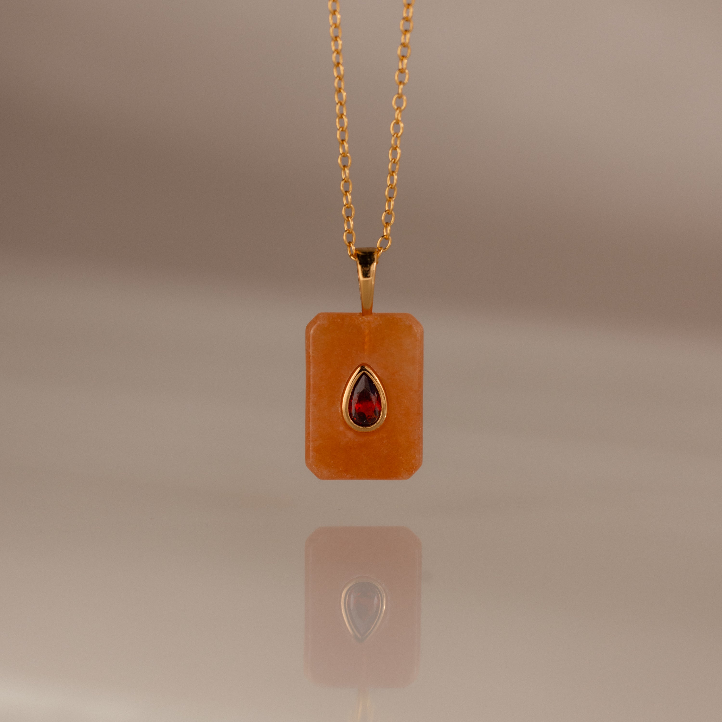 The Garnet Crystal Necklace features a gold chain with a rectangular orange pendant and a teardrop red gemstone, making it a unique birthstone jewelry piece or an eye-catching Birthstone Crystal Necklace.