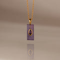 Gold chain necklace with a rectangular purple pendant and gold accent, reflected on a smooth surface—an elegant Amethyst Crystal Necklace perfect for adding a touch of birthstone jewelry to any look.