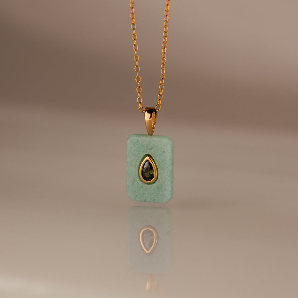 The Peridot Crystal Necklace features a rectangular green crystal pendant in a gold bezel setting, highlighted by a gold-trimmed teardrop accent at the center.