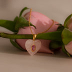 Birthstone Crystal Necklace featuring a gold chain with a pink heart-shaped pendant, displayed on a pink rose over a soft beige background.