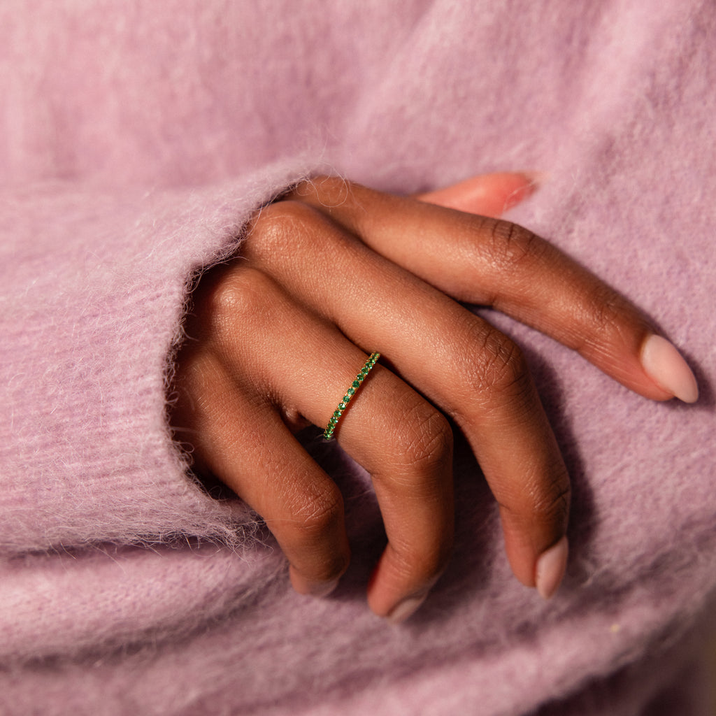 A hand adorned with the Endless Eternity Birthstone Ring, featuring a green gemstone, rests on a lavender fuzzy sweater; nails are painted pale pink.
