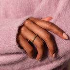 A hand adorned with the Endless Eternity Birthstone Ring, featuring a green gemstone, rests on a lavender fuzzy sweater; nails are painted pale pink.