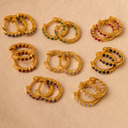 Nine pairs of gold stacking earrings, including Inside Out Opal Huggies with colorful gemstones, arranged on a beige background.